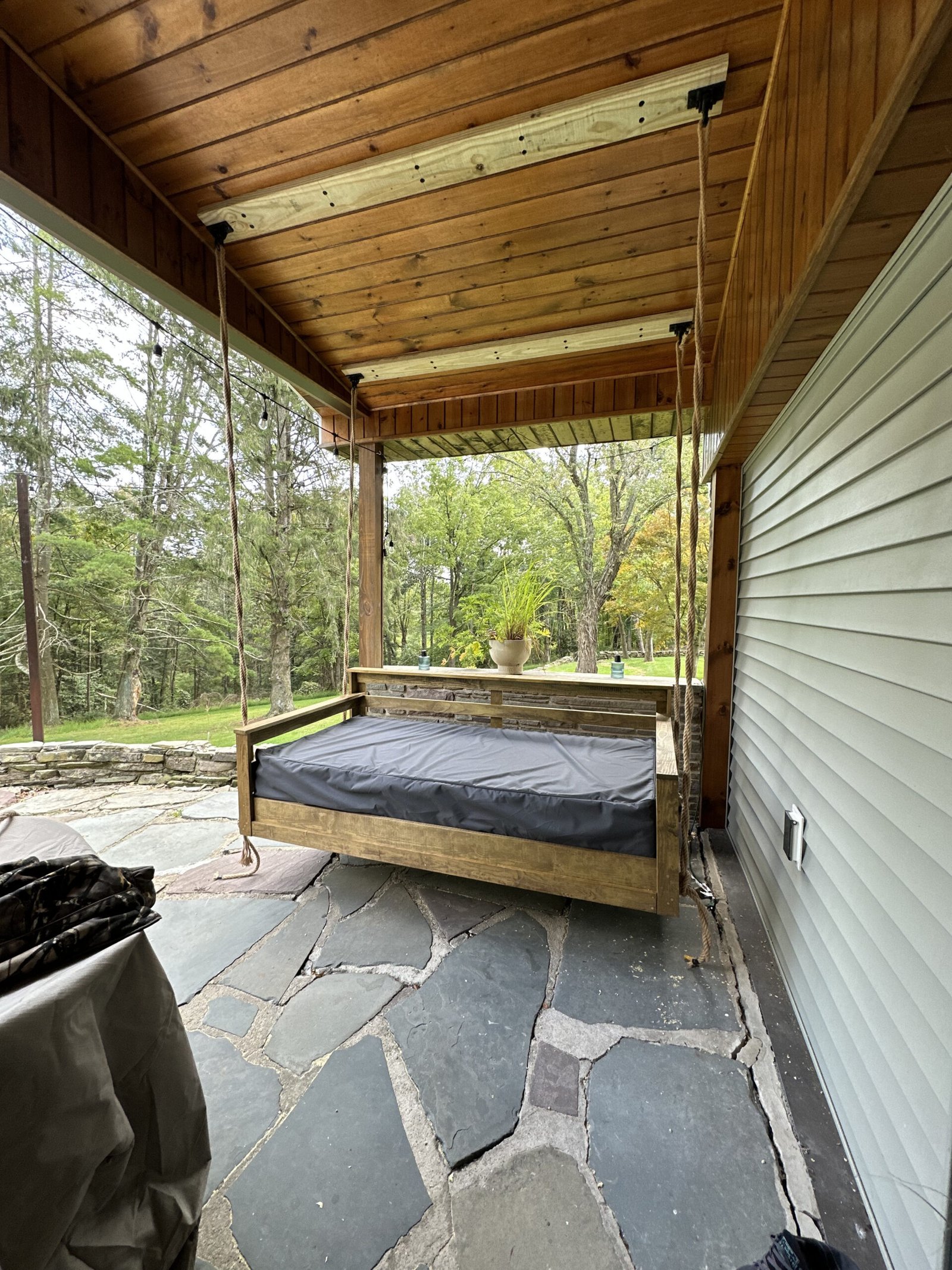 Wooden swing bed on a stone patio under a covered porch, surrounded by trees, emphasizing outdoor living and custom deck features.