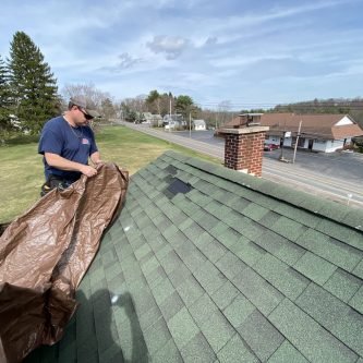 Worker preparing a tarp on a green shingle roof during a roofing project in Wilkes-Barre, Pennsylvania, with a residential area in the background.