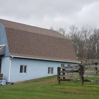 Barn-style building with a new roof, featuring blue siding and surrounded by a grassy area and wooden fence, reflecting E-Stern Construction's recent project in Wilkes-Barre, PA.